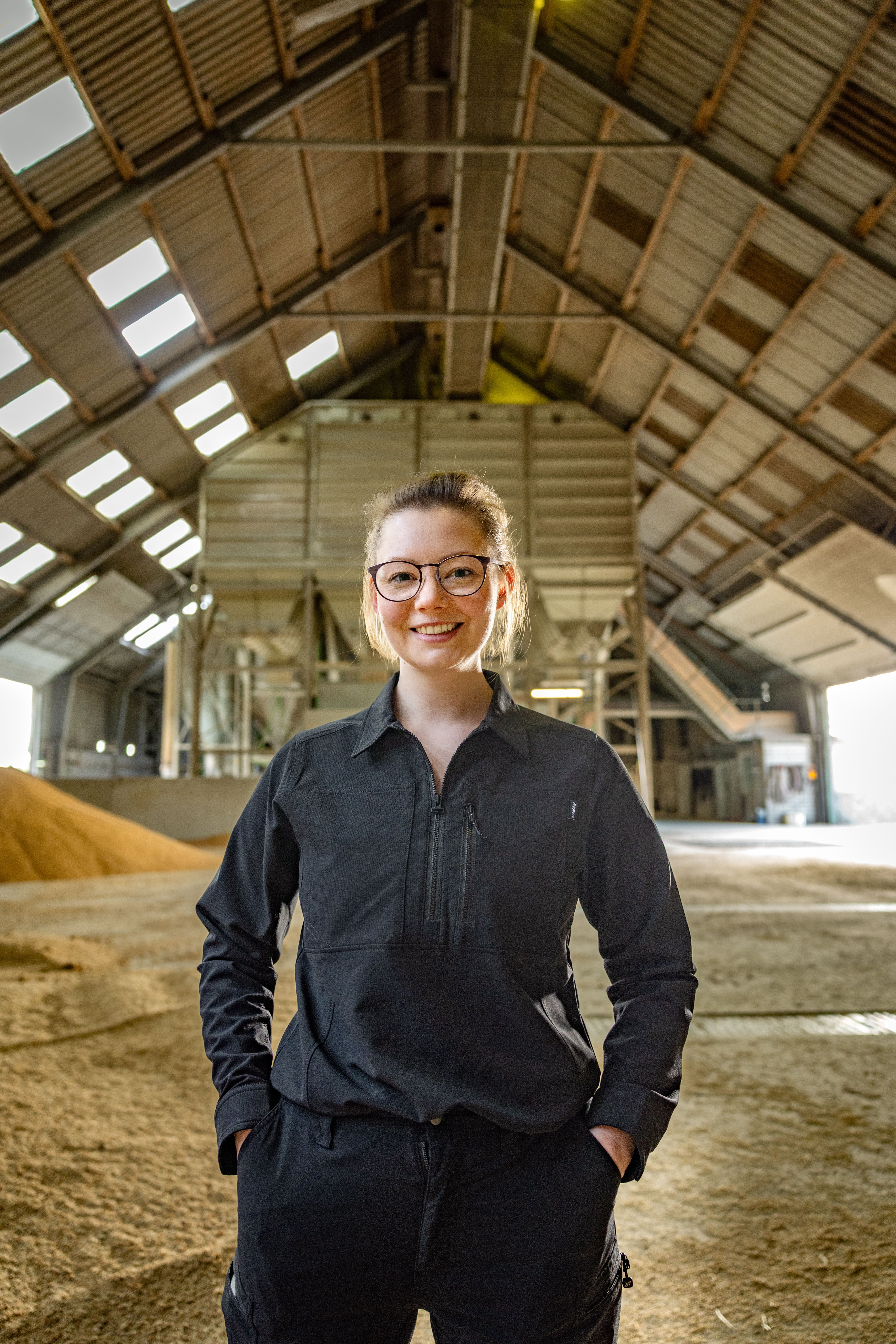 Jenna Struck steht in einer großen, lichtdurchfluteten Halle mit hohem, schrägem Dach. Im Hintergrund sind industrielle Anlagen und Schüttgut zu sehen. Sie trägt eine schwarze, langärmelige Arbeitsjacke und eine dunkle Hose, die Hände in den Hosentaschen. Das Licht fällt durch die Fenster im Dach und erzeugt helle Flächen auf dem Boden.